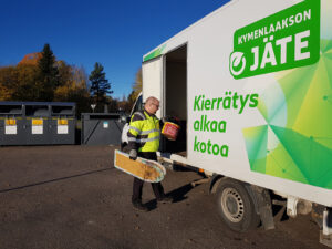 Jätehuollon työntekijä lastaa kierrätettäviä roskia Kymenlaakson Jätteen logolla varustettuun autoon aurinkoisena päivänä.